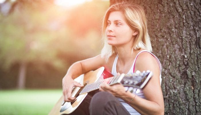 Garota loira tocando violão embaixo de uma árvore