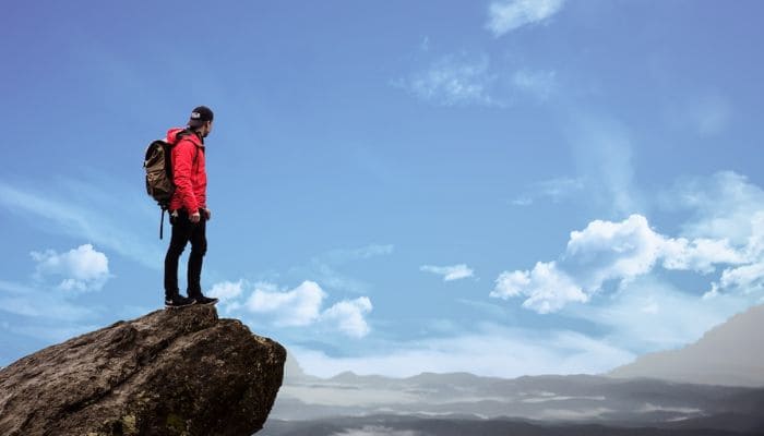 Homem com traje de alpinismo no topo de um rochedo olhando para o horizonte