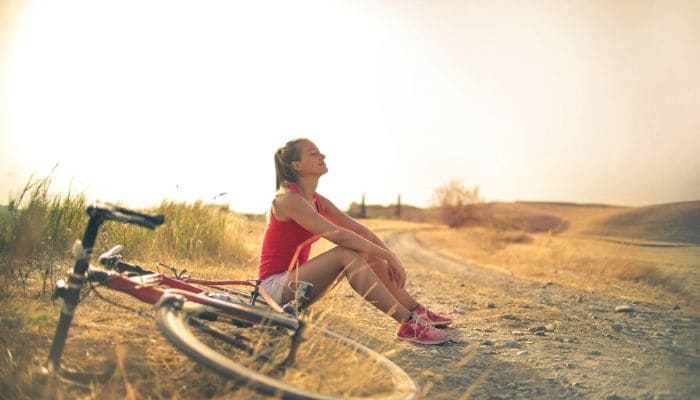 Mulher com semblante feliz sentada no chão de um campo ao lado de uma bicicleta