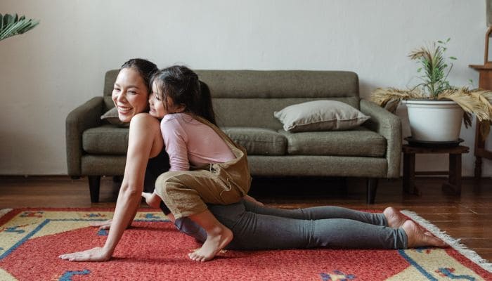 Mulher sorridente fazendo alongamento no tapete da sala com sua filha nas costas