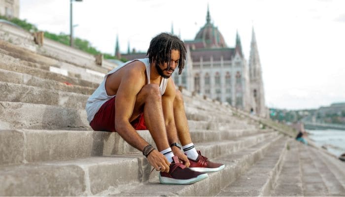 Homem de barba e cablo dread look com roupas de treino amarrando cadarço do tênis sentado em uma escadaria