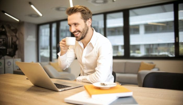 Homem sorridente tomando café enquanto olha para a tela do notebook