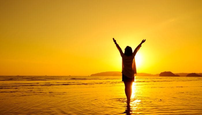 Mulher em uma praia com os braços levantados aos ceus agradecendo ao nascer do sol