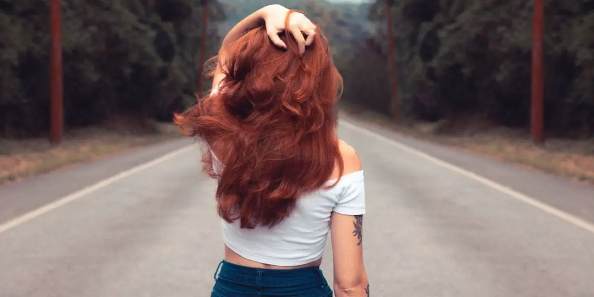 Virtudes: Bela mulher ruiva de costas no meio de uma estrada com a mão nos cabelos.