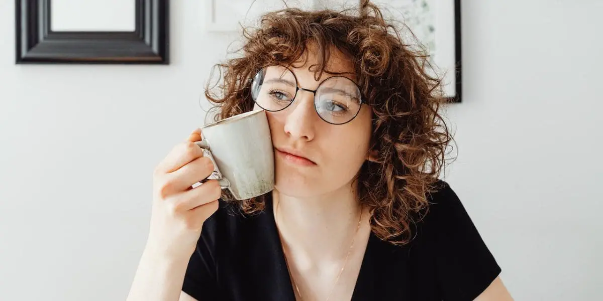 Pensamentos e emoções: Garota ruiva de óculos com semblante desanimado com uma caneca encostada na bochecha.