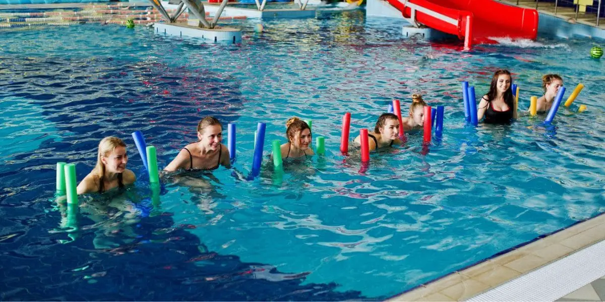 Grupo de mulheres na piscina usando espaguetes flutuadores na aula de hidroginástica.