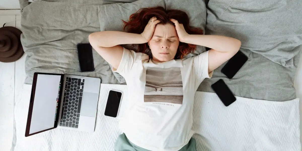 Zona de conforto: Mulher deitada na cama com as mãos na cabeça ao lado de um notebook e quatro celulares.