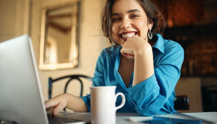 moça-alegre-desfrutando-do-home-office-sentada-mesa-usando-computador-portátil-bebendo-café-sorrindo